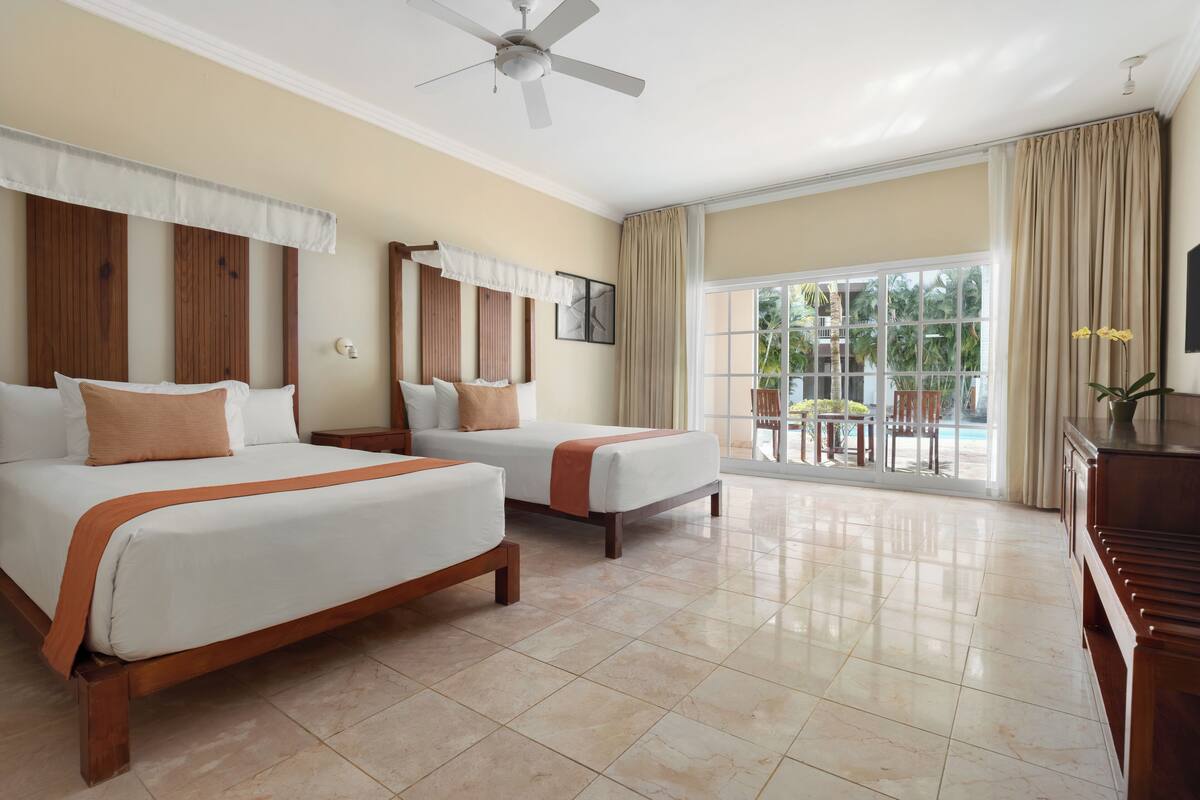 Habitación de hotel con dos camas, ventilador de techo, ventana grande con vistas al exterior y balcón con mesa y sillas.