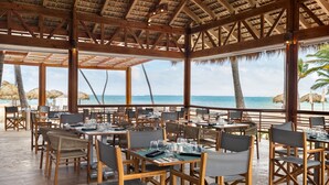 An outdoor dining area with wooden tables and chairs, thatched umbrellas, and a view of the ocean.