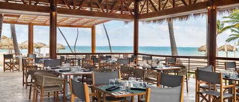 An outdoor dining area with wooden tables and chairs, thatched umbrellas, and a view of the ocean.