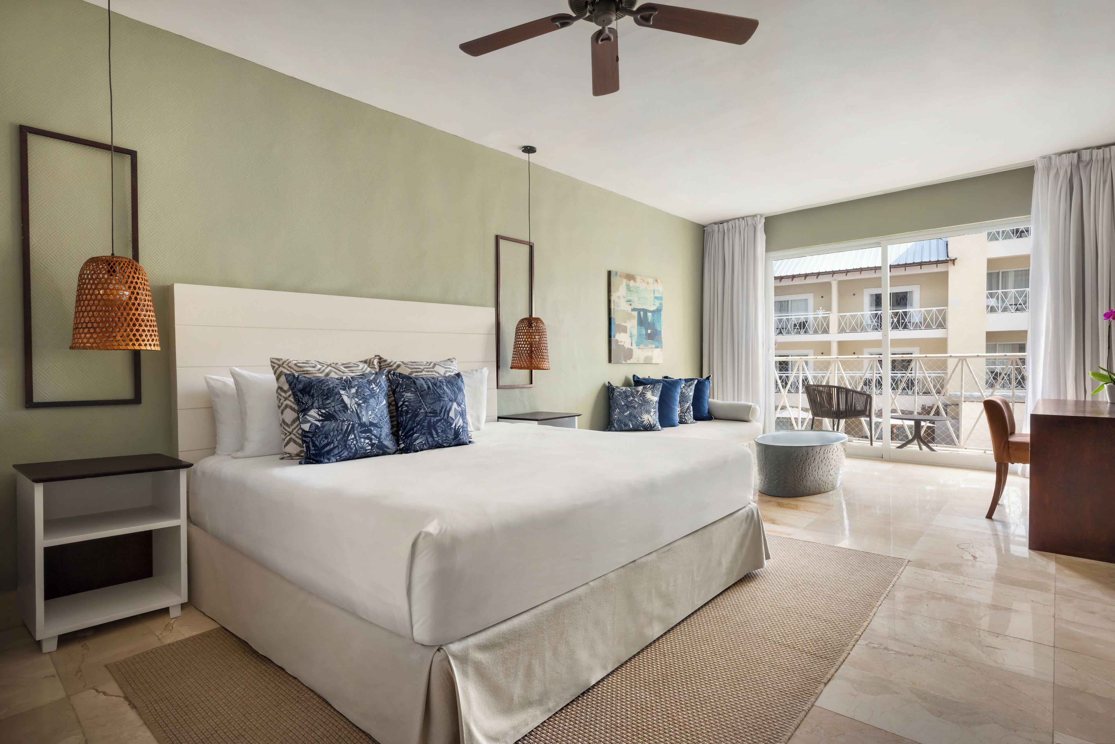 Habitación de hotel con una cama grande, ventilador de techo, balcón con vista al edificio y un escritorio con silla.