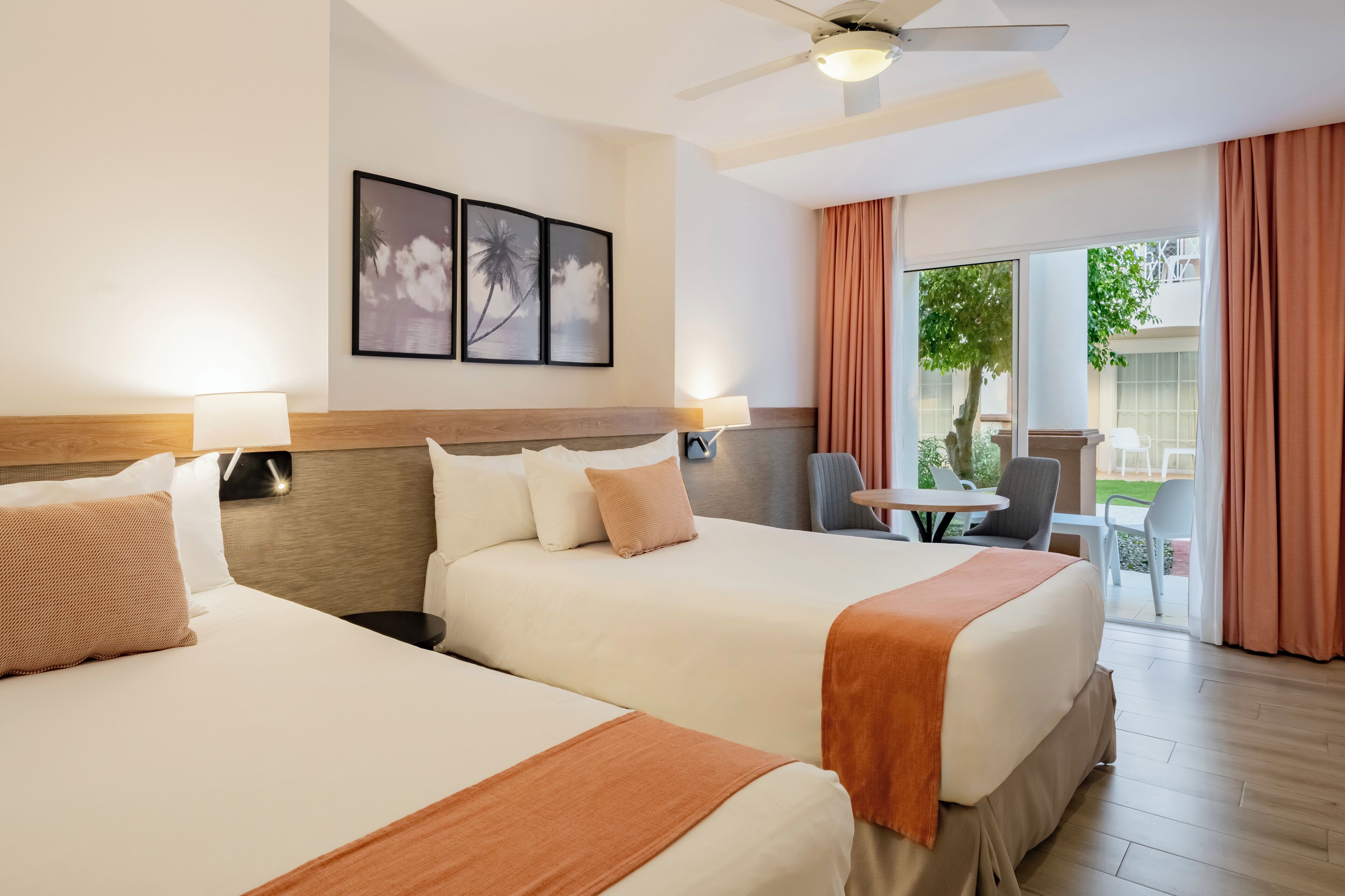 A hotel room with two beds, a ceiling fan, a table with chairs, and a view of the outdoors.