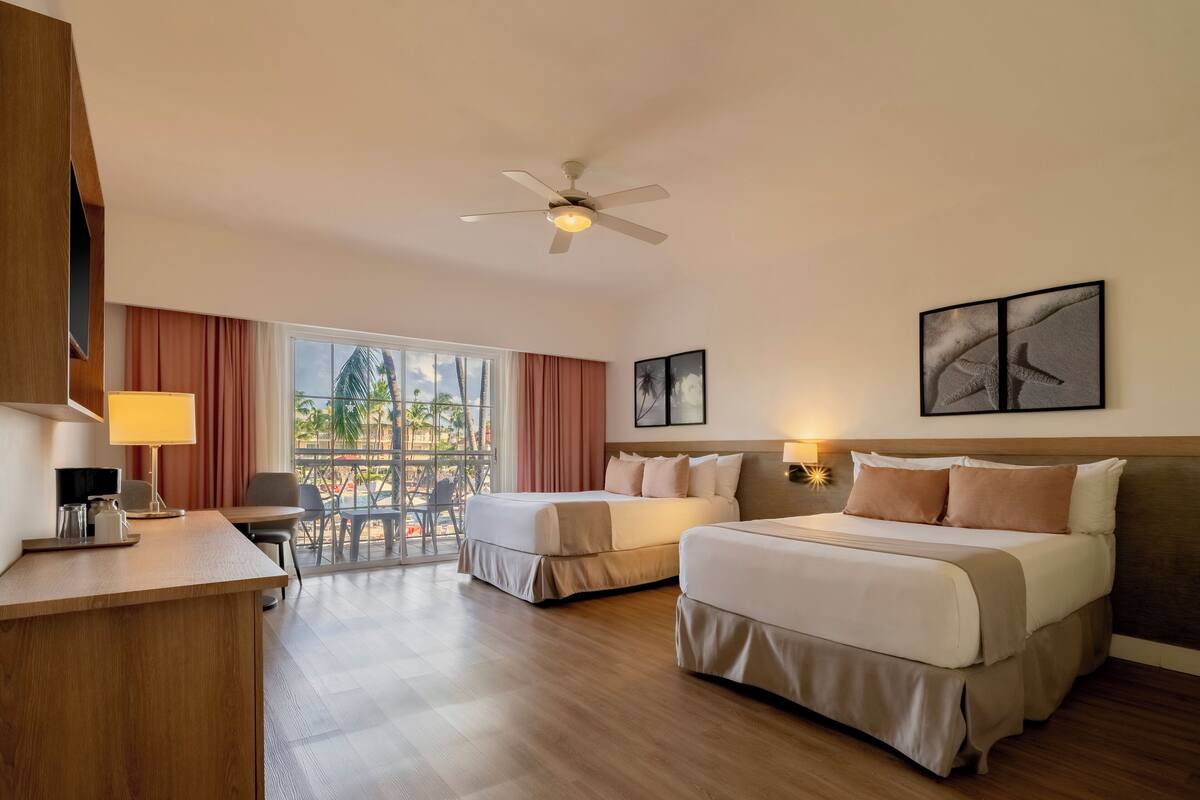 Habitación de hotel con dos camas, un escritorio, una silla, un ventilador de techo y un amplio ventanal con vistas.