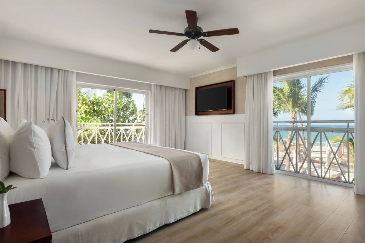 Un dormitorio con una cama grande, un ventilador de techo, un televisor montado en la pared y un balcón con vistas a la playa.