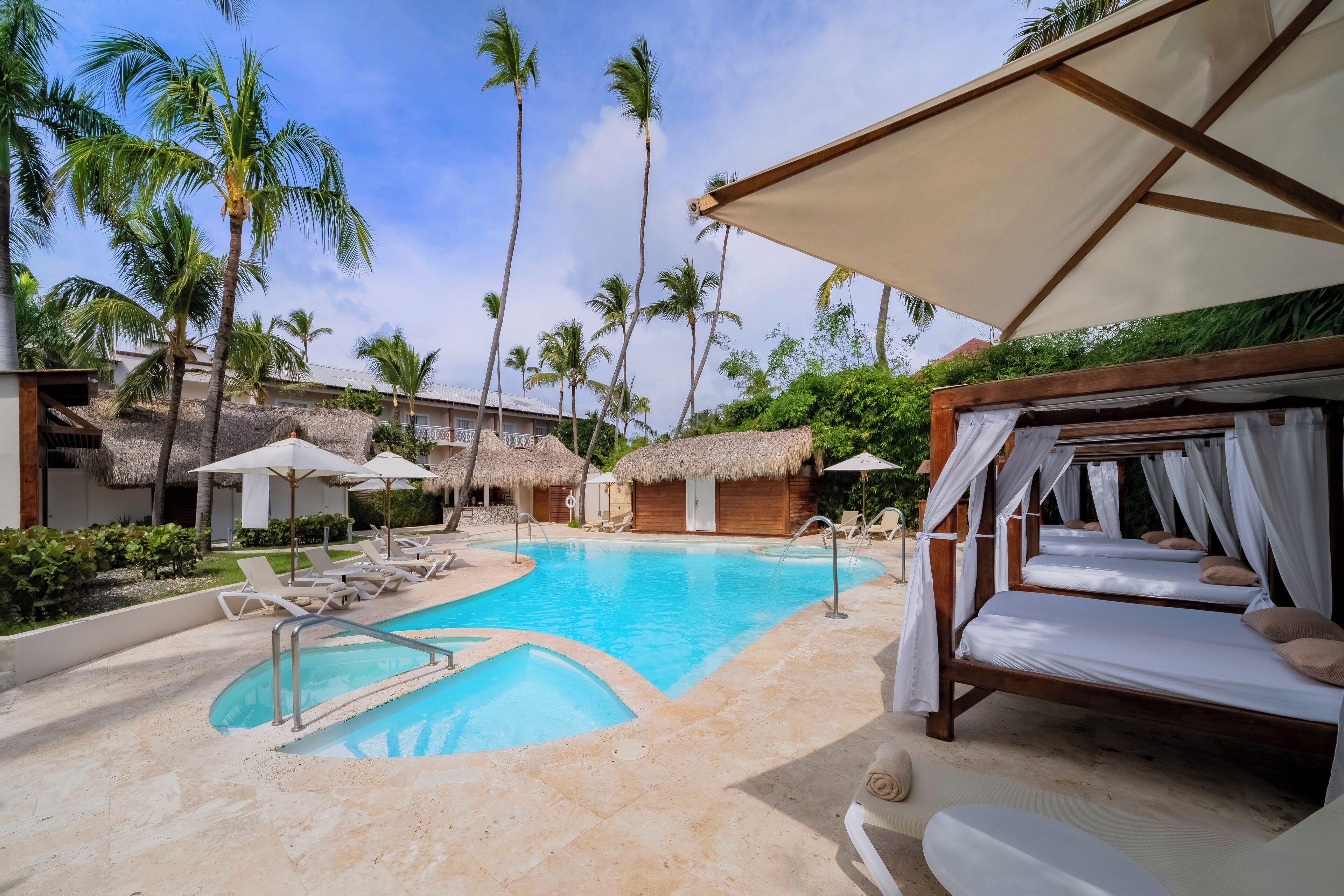 A resort pool area with a clear blue pool, lounge chairs, and thatched-roof structures.