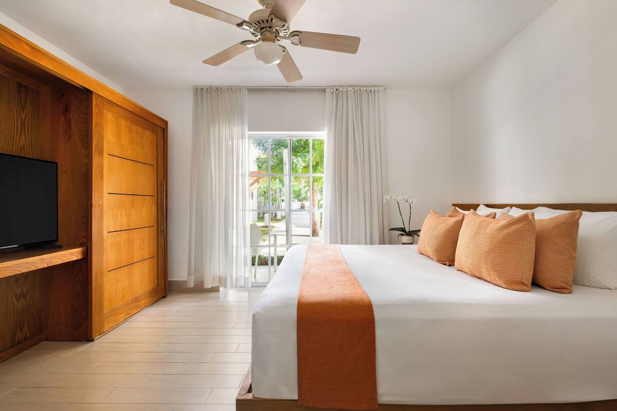Habitación de hotel con cama, televisión, ventilador de techo y ventana con cortinas.