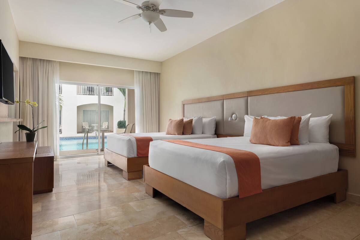Habitación de hotel con dos camas, ventilador de techo, televisor y vista a una piscina.
