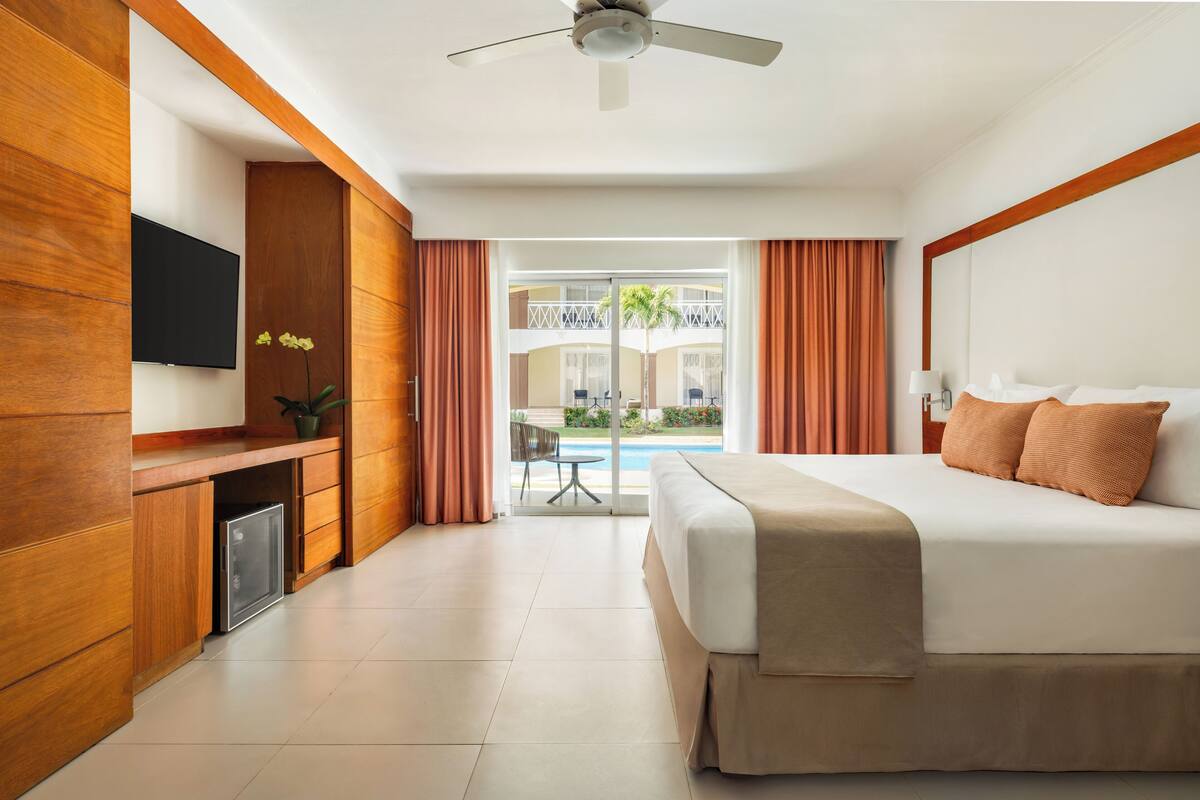 Una habitación de hotel con cama, televisión, escritorio con computadora, planta y vista a una piscina y palmeras.