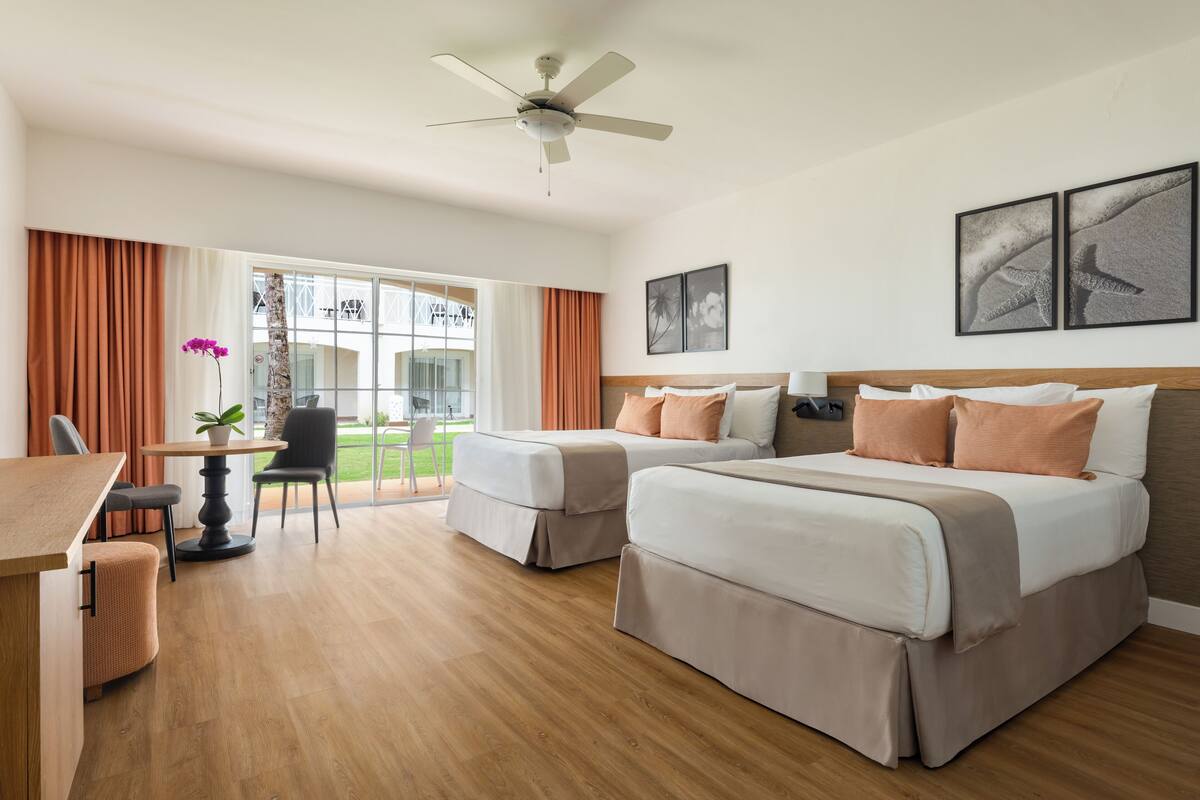 Habitación de hotel con dos camas, un escritorio, una silla, una mesa con una planta y un balcón con vistas.