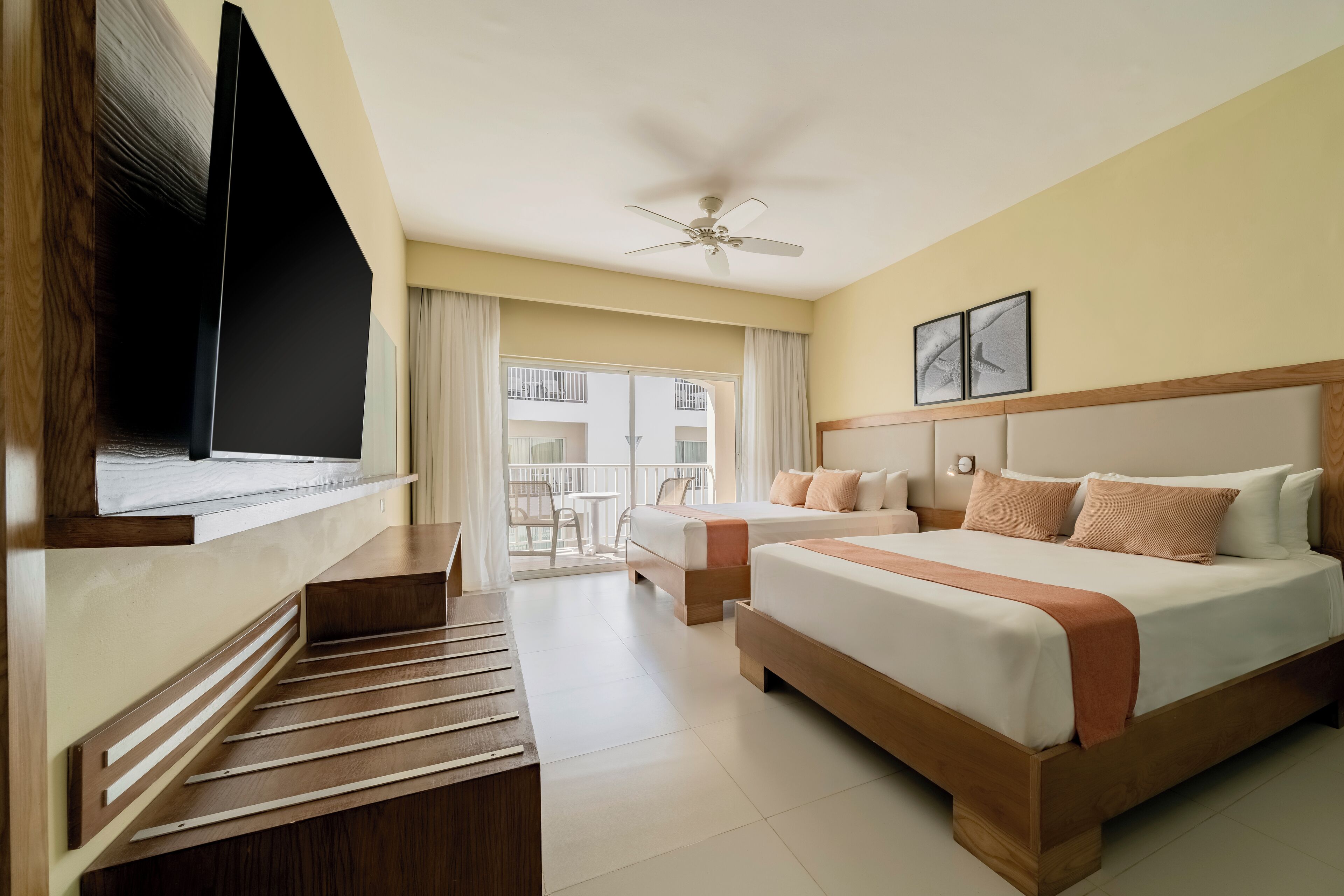 A hotel room with a bed, a TV, a ceiling fan, and a balcony with a table and chairs.
