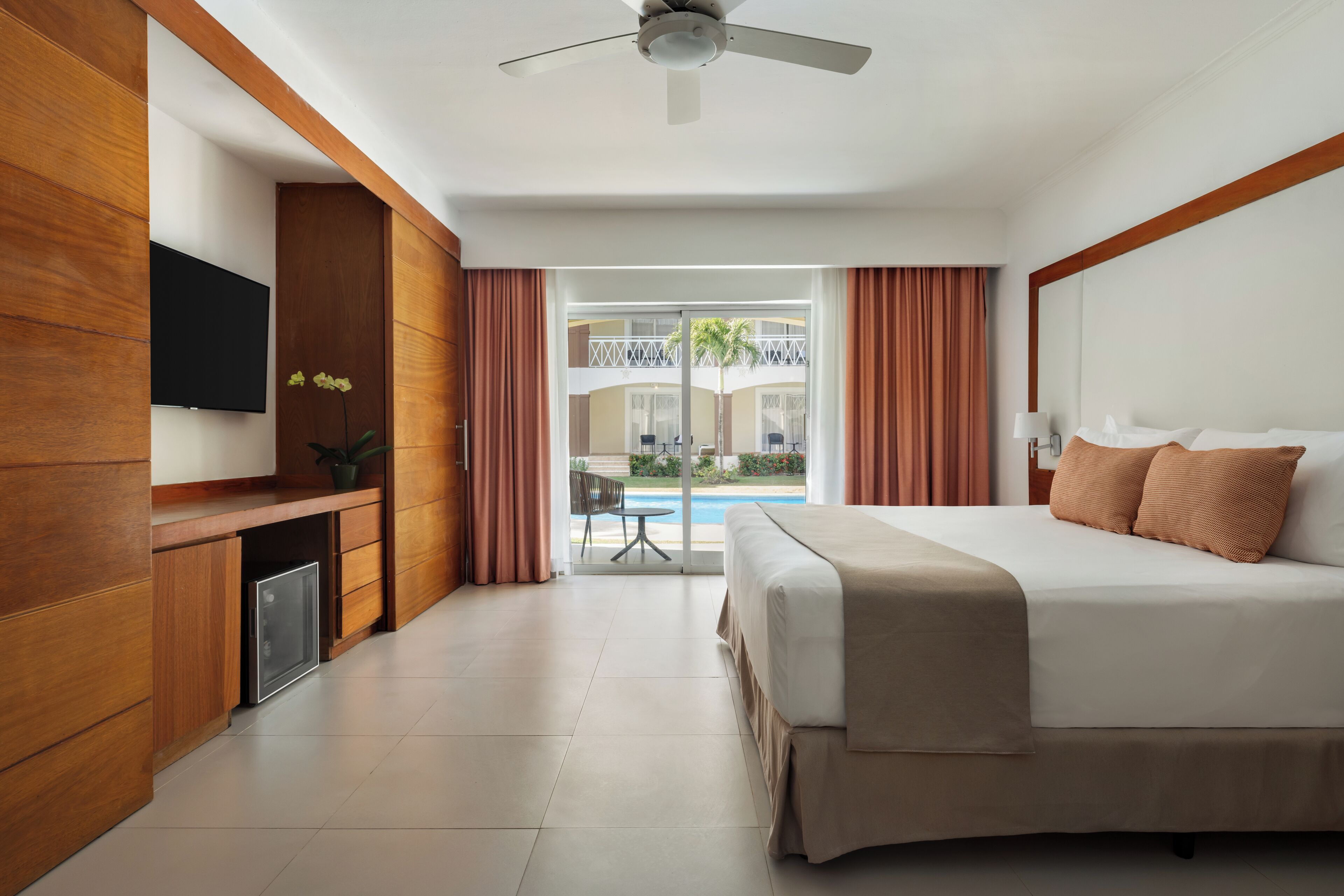 A hotel room with a bed, a TV, a desk, and a view of a pool and building.