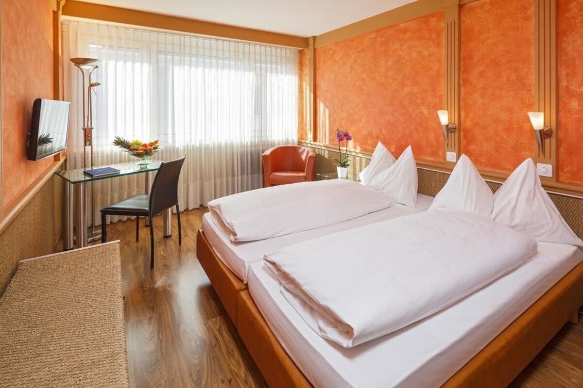 A hotel room with two beds, a desk, a chair, a small table with fruit, and a window with curtains.