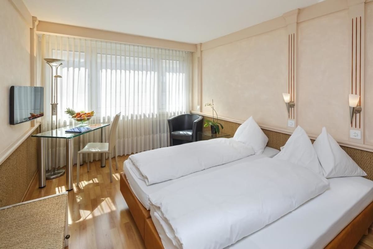 A hotel room with two beds, a desk, a chair, a TV, and a window with blinds.