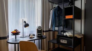Une chambre d’hôtel compacte comprenant un petit bureau, une machine à café, un panier de fruits, une lampe, une chaise, une étagère suspendue avec des vêtements et un lit superposé avec une couverture.