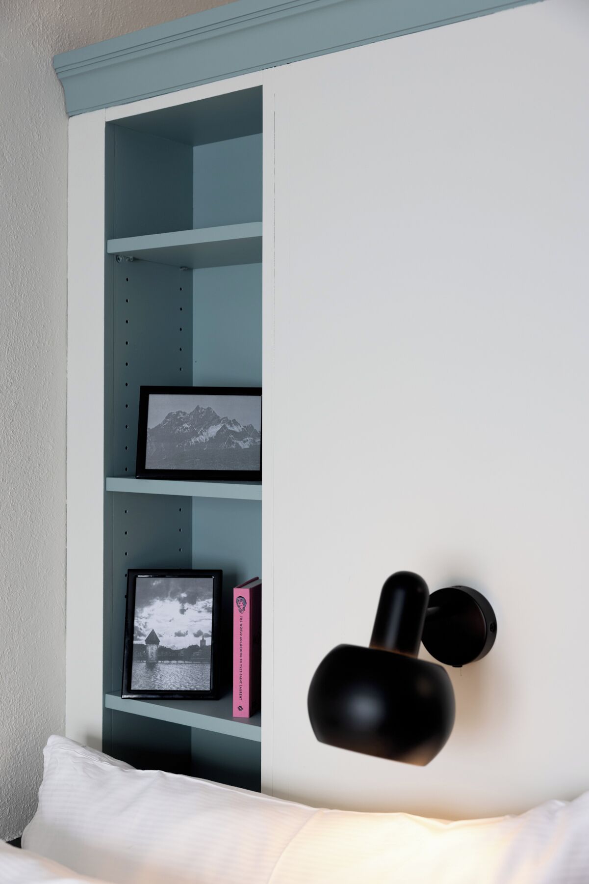 A bedroom with a bed, a wall-mounted shelf with framed pictures, and a black object on the wall.