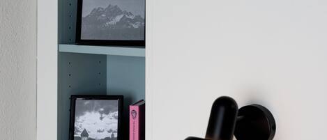 A bedroom with a bed, a wall-mounted shelf with framed pictures, and a black object on the wall.