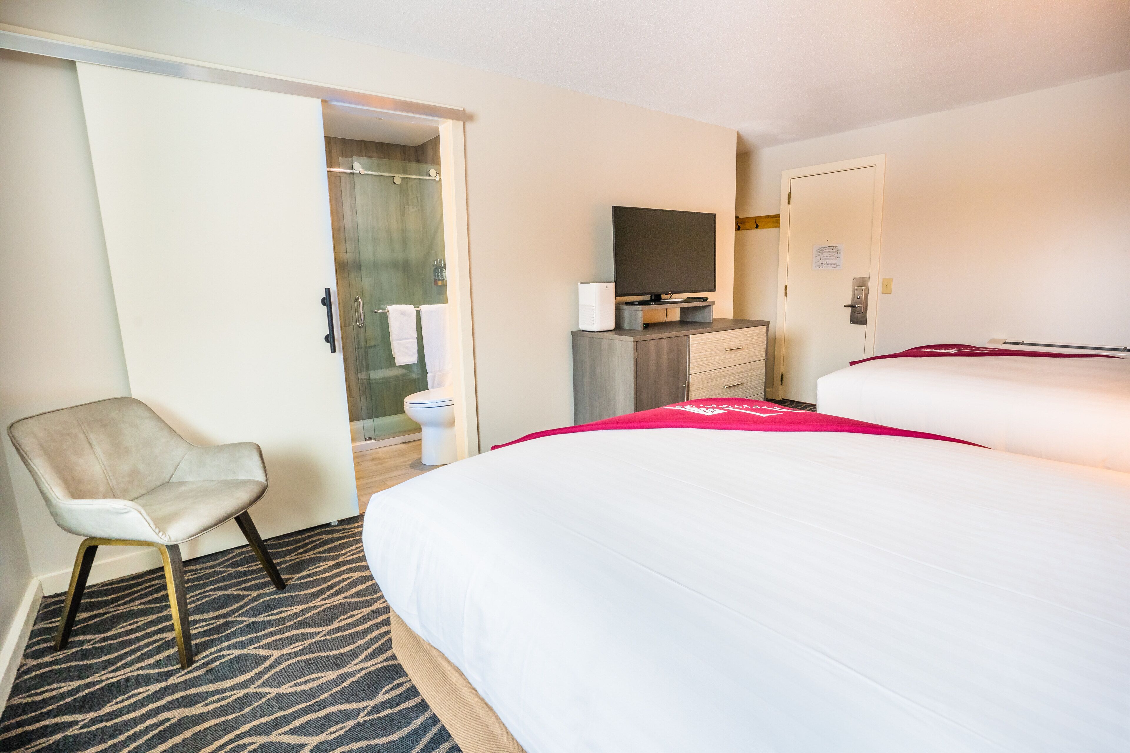A hotel room with a bed, a chair, a desk with a TV, and a bathroom visible through an open door.