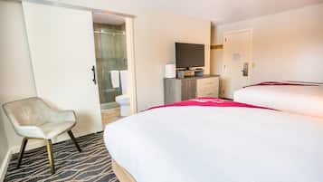 Une chambre d’hôtel avec un lit, une chaise, un bureau avec une télévision et une salle de bain visible à travers une porte ouverte.