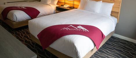 A hotel room with two beds, each with a wooden headboard, white bedding, and a red blanket with the hotel logo. There are two bedside lamps and a painting on the wall above the beds.