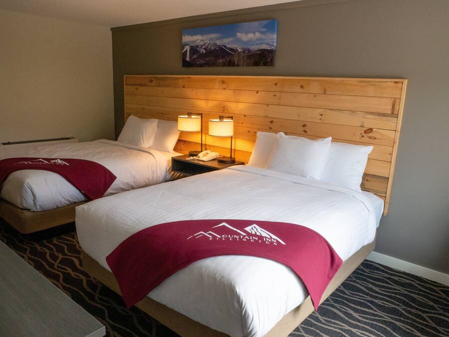 Habitación de hotel con dos camas, cada una con cabecero de madera, ropa de cama blanca y una manta roja con el logotipo del hotel. Hay dos lámparas de noche y un cuadro en la pared sobre las camas.