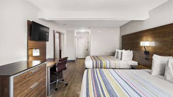 A hotel room with a wooden desk, a television, two beds with striped bedding, a chair, and a lamp.