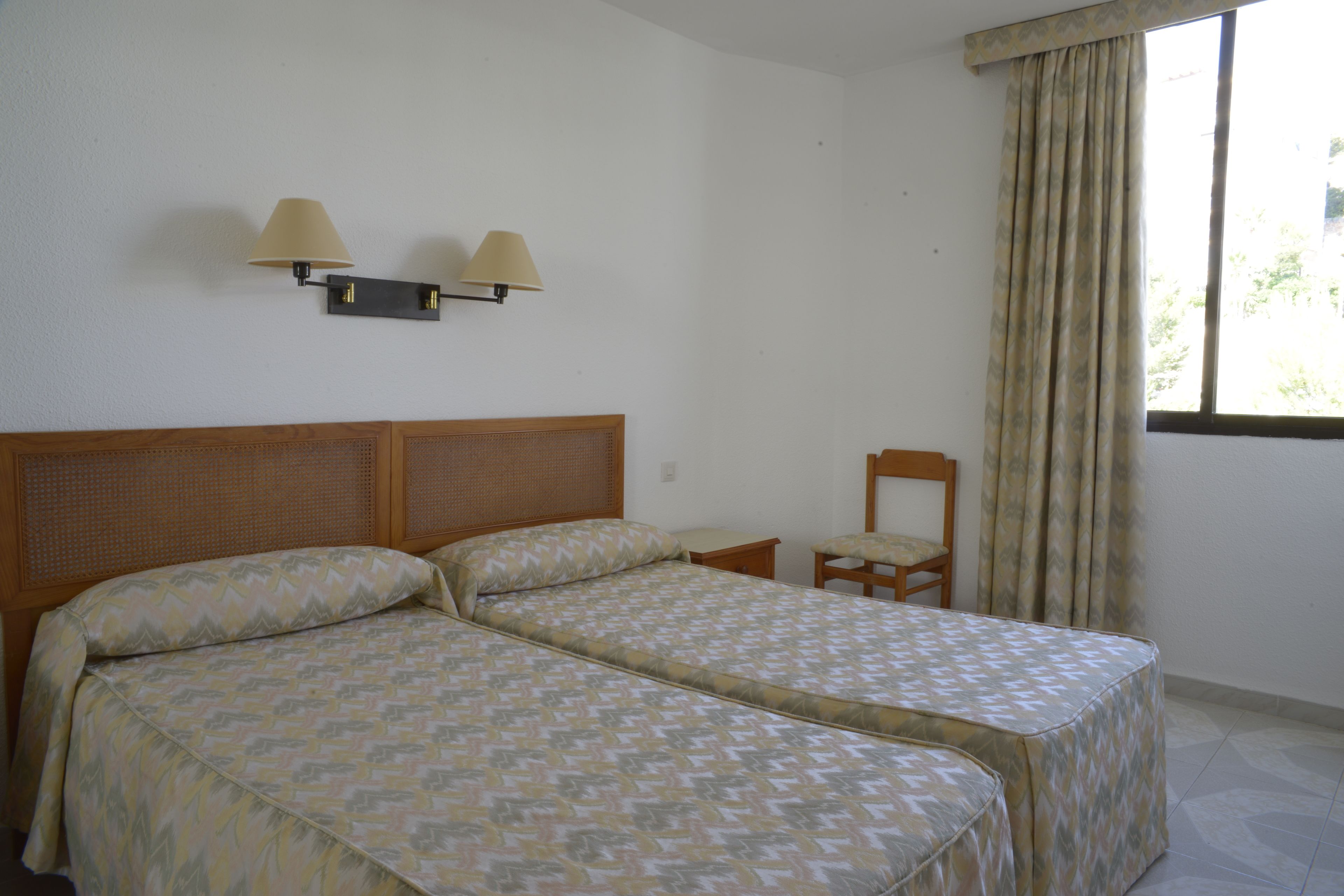 A double bed with a patterned bedspread, two wall-mounted lamps, a wooden chair, and a window with curtains.
