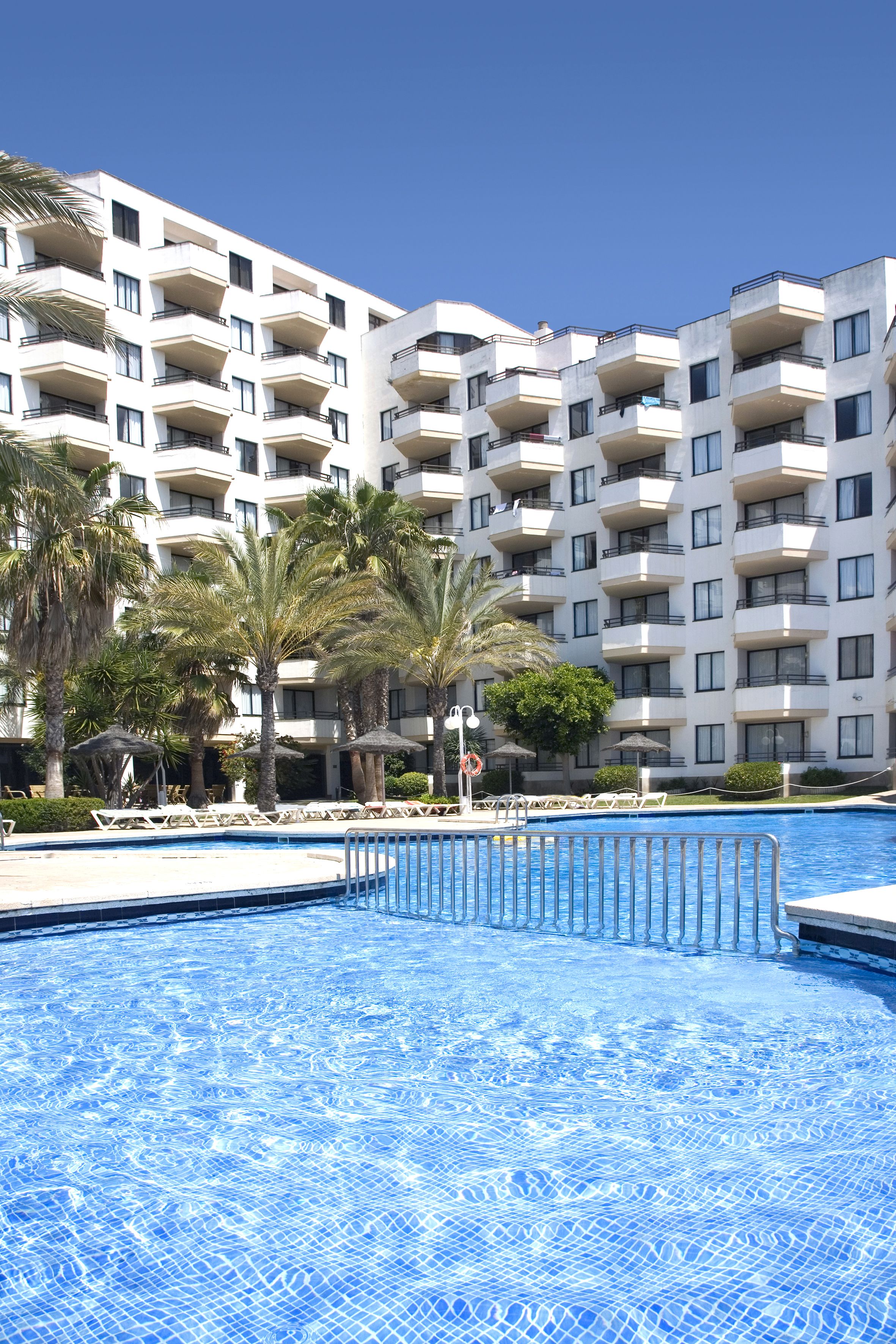 A swimming pool with a clear blue water surface, surrounded by lounge chairs and palm trees. In the background, there is a multi-story building with balconies.