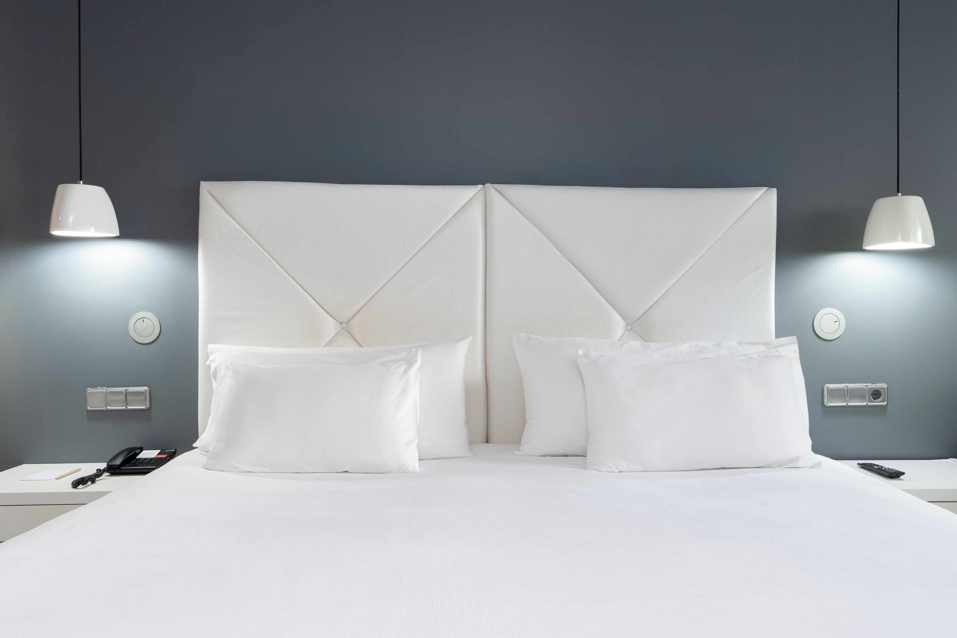 A neatly made bed with white linens, two bedside tables with a phone and a remote control, and two pendant lights on the wall.
