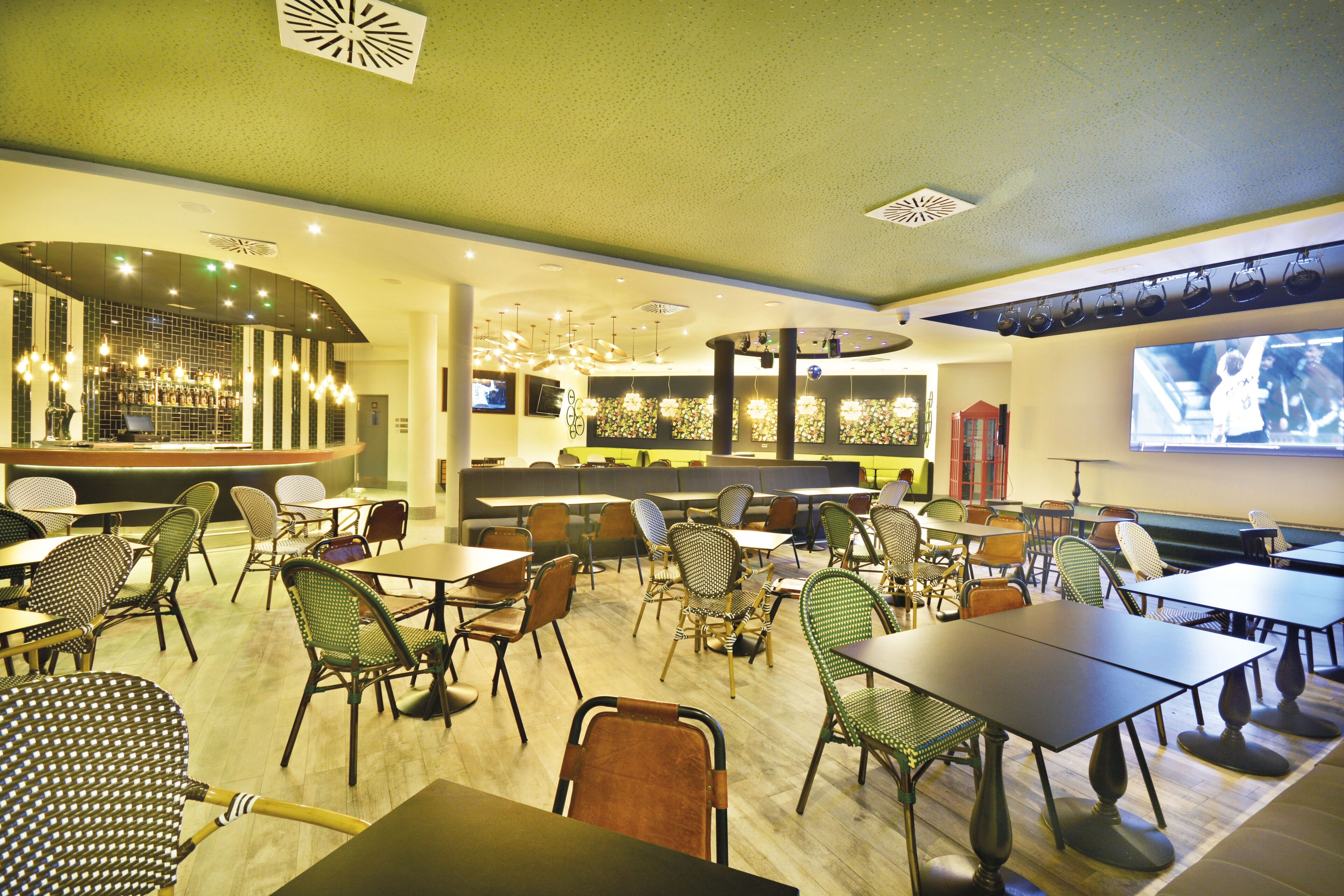 A restaurant with multiple tables and chairs, a bar area, and a television screen displaying a program.