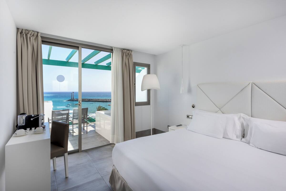 A hotel room with a bed, a desk with a coffee maker, a chair, and a view of the ocean through large windows.