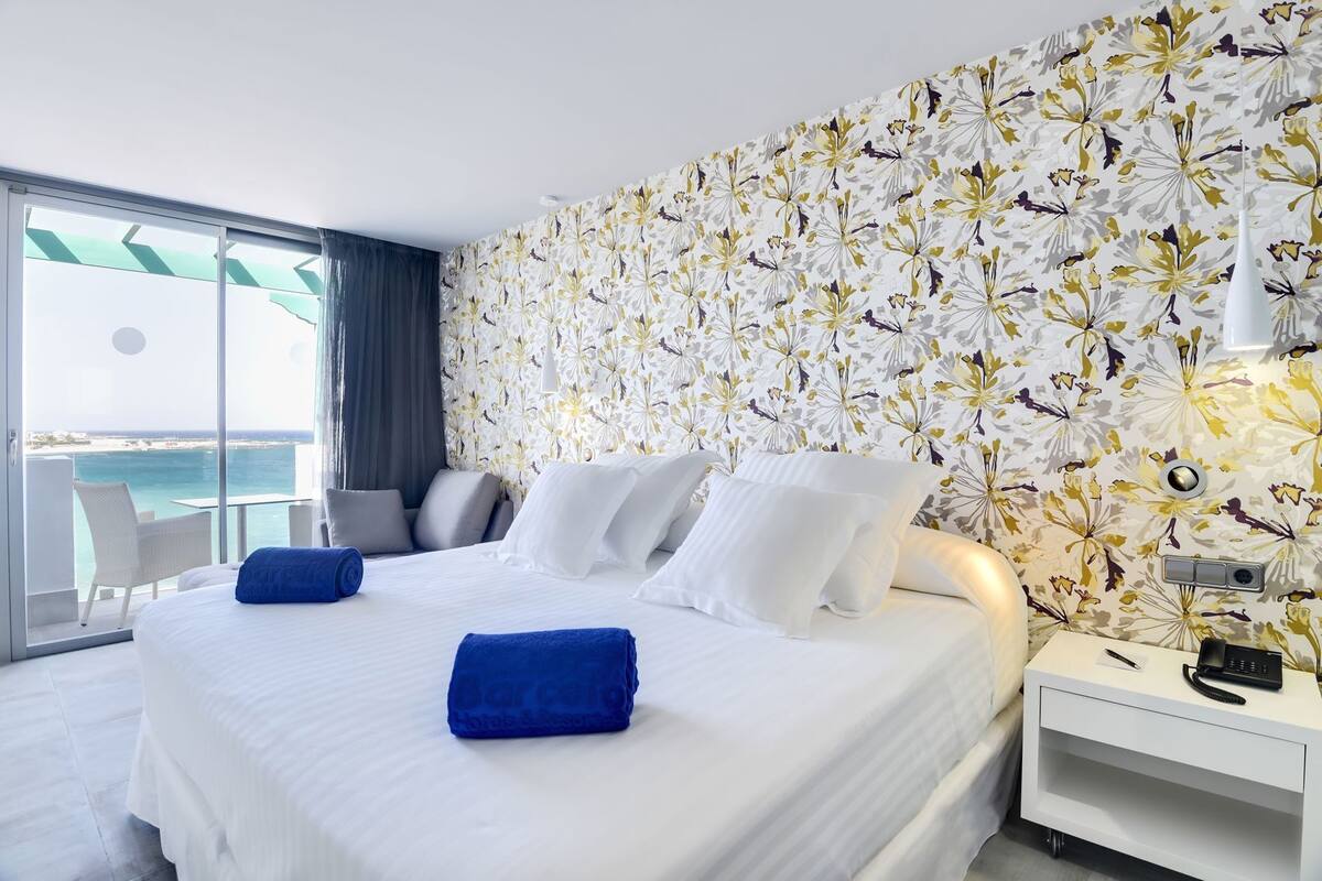A hotel room with a large bed, a nightstand, a chair, and a view of the ocean.
