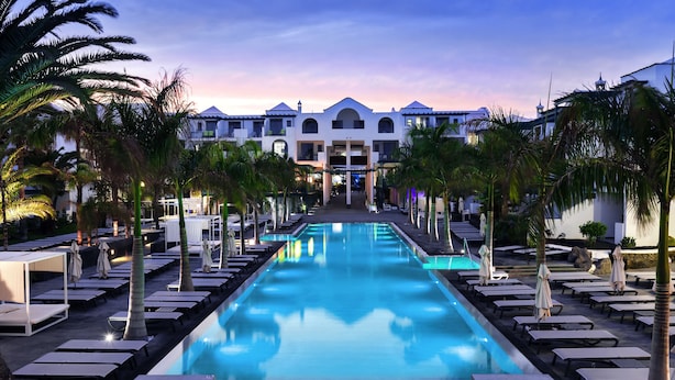 A pool area with lounge chairs and umbrellas, surrounded by palm trees, leading to a building with multiple balconies. - Barceló Teguise Beach - Adults only (Teguise)