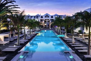 A pool area with lounge chairs and umbrellas, surrounded by palm trees, leading to a building with multiple balconies. - Barceló Teguise Beach - Adults only (Teguise)