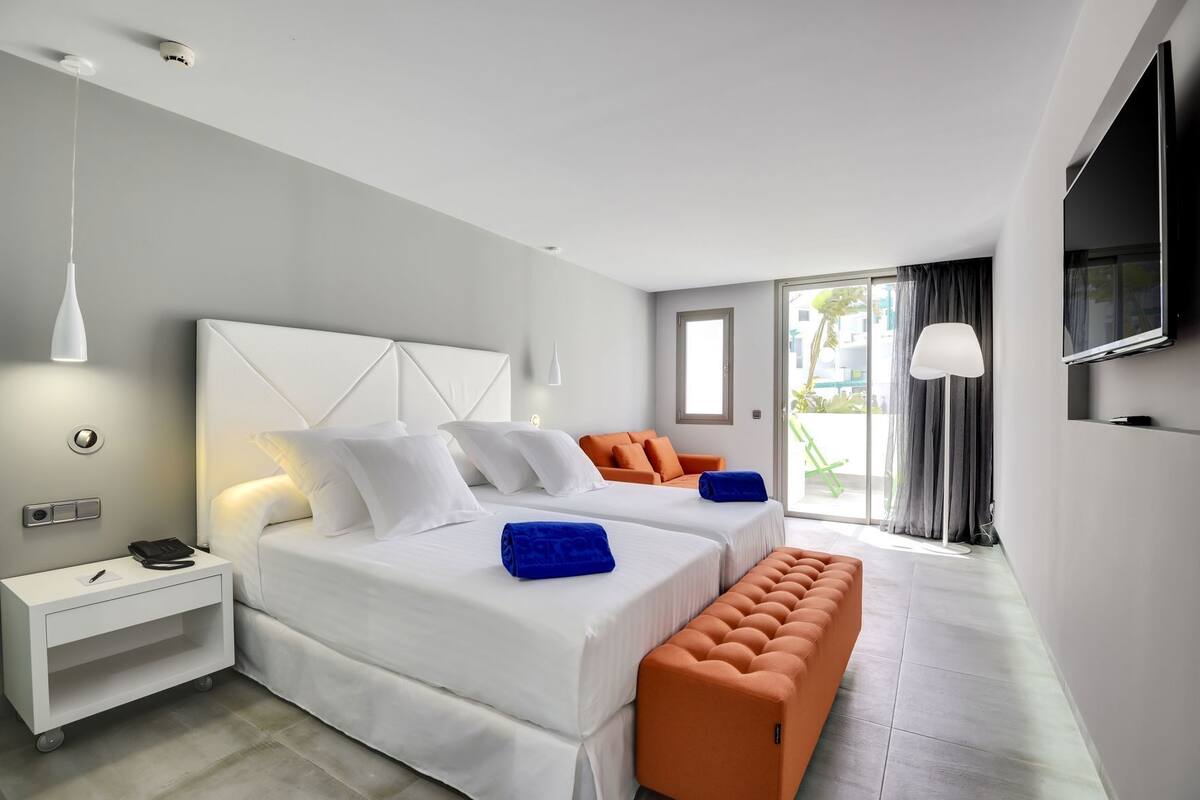 A modern hotel room with two beds, a bench, a TV, and a balcony.