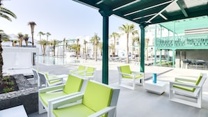 A covered outdoor seating area with white and green lounge chairs, a pool, and a view of palm trees and buildings. - Barceló Teguise Beach - Adults only (Teguise)