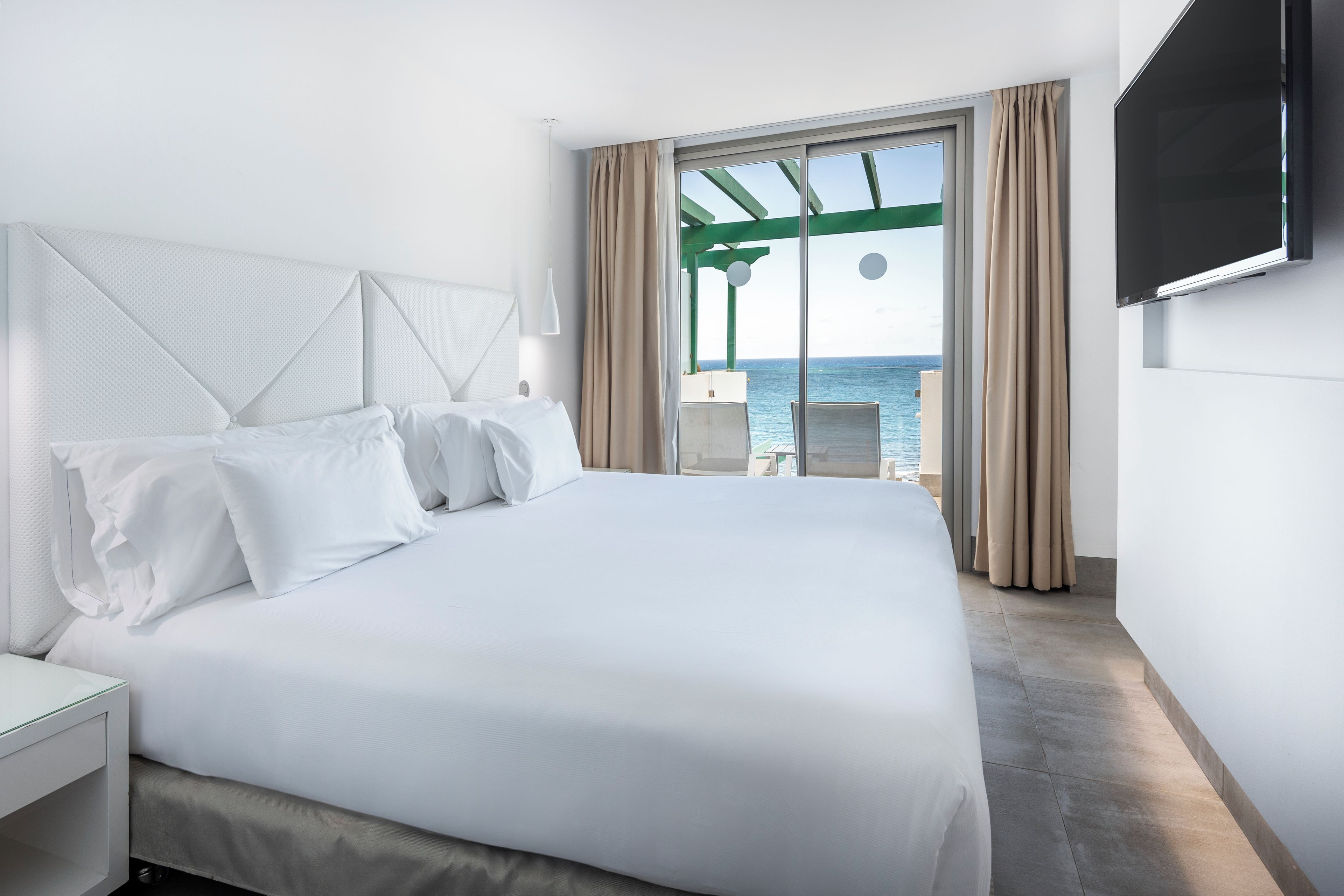 Habitación de hotel con una cama grande, un televisor montado en la pared y vista al mar por la ventana.