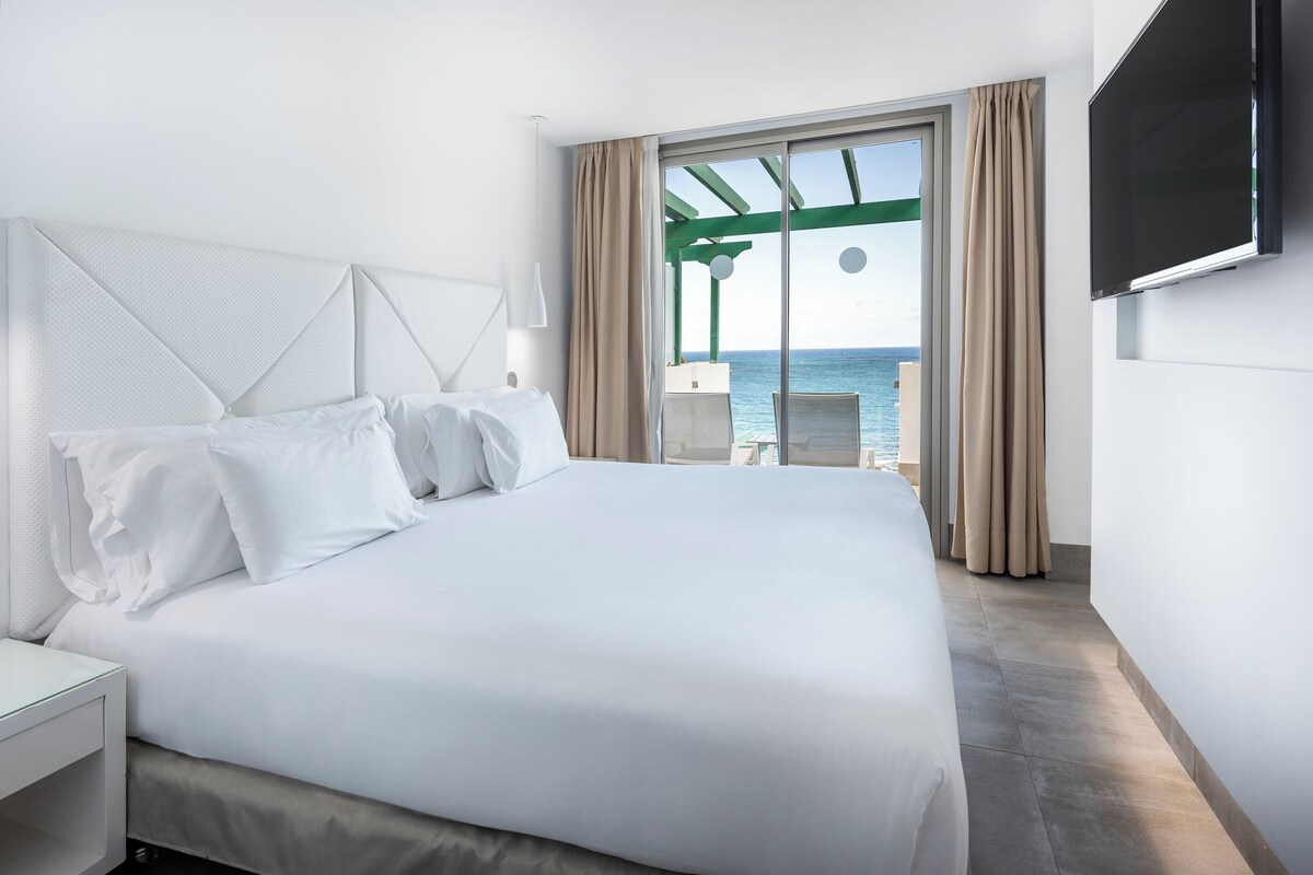 Habitación de hotel con una cama grande, un televisor montado en la pared y vista al mar por la ventana.