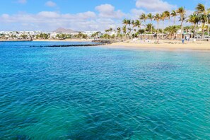 Escena de playa costera con aguas turquesas cristalinas, palmeras y edificios al fondo.