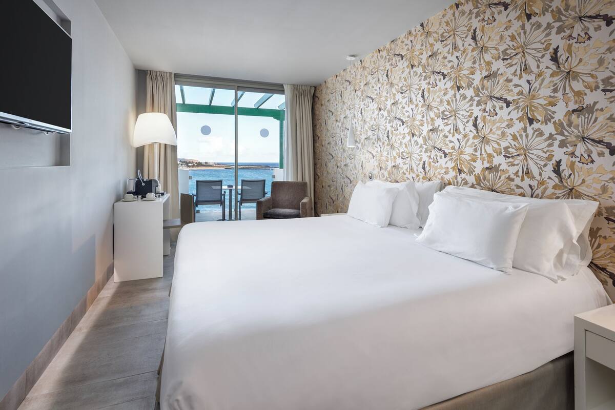 A hotel room with a large bed, a desk with a lamp, a chair, and a view of the sea through a large window.