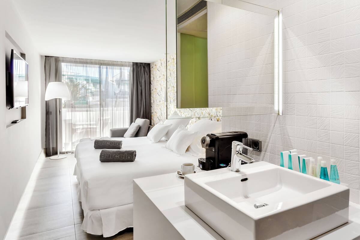 A hotel room with a bed, a bedside table with a coffee maker, a large mirror, and a bathroom with a sink and toiletries.