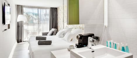 A hotel room with a bed, a bedside table with a coffee maker, a large mirror, and a bathroom with a sink and toiletries.