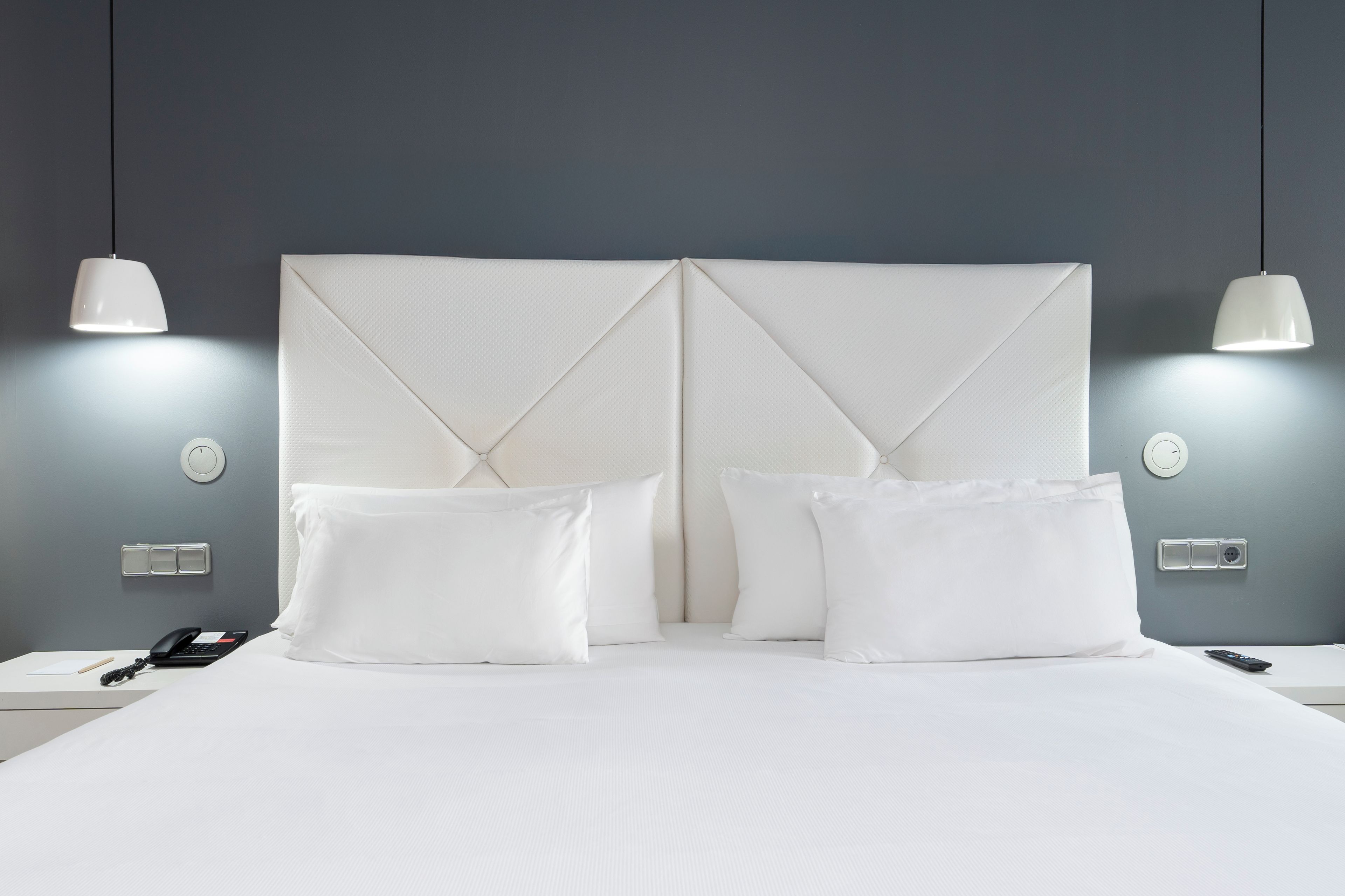 Ein ordentlich bezogenes Bett mit weißen Leinentüchern, zwei Nachttische mit Telefon und Fernbedienung sowie zwei Pendelleuchten an der Wand.