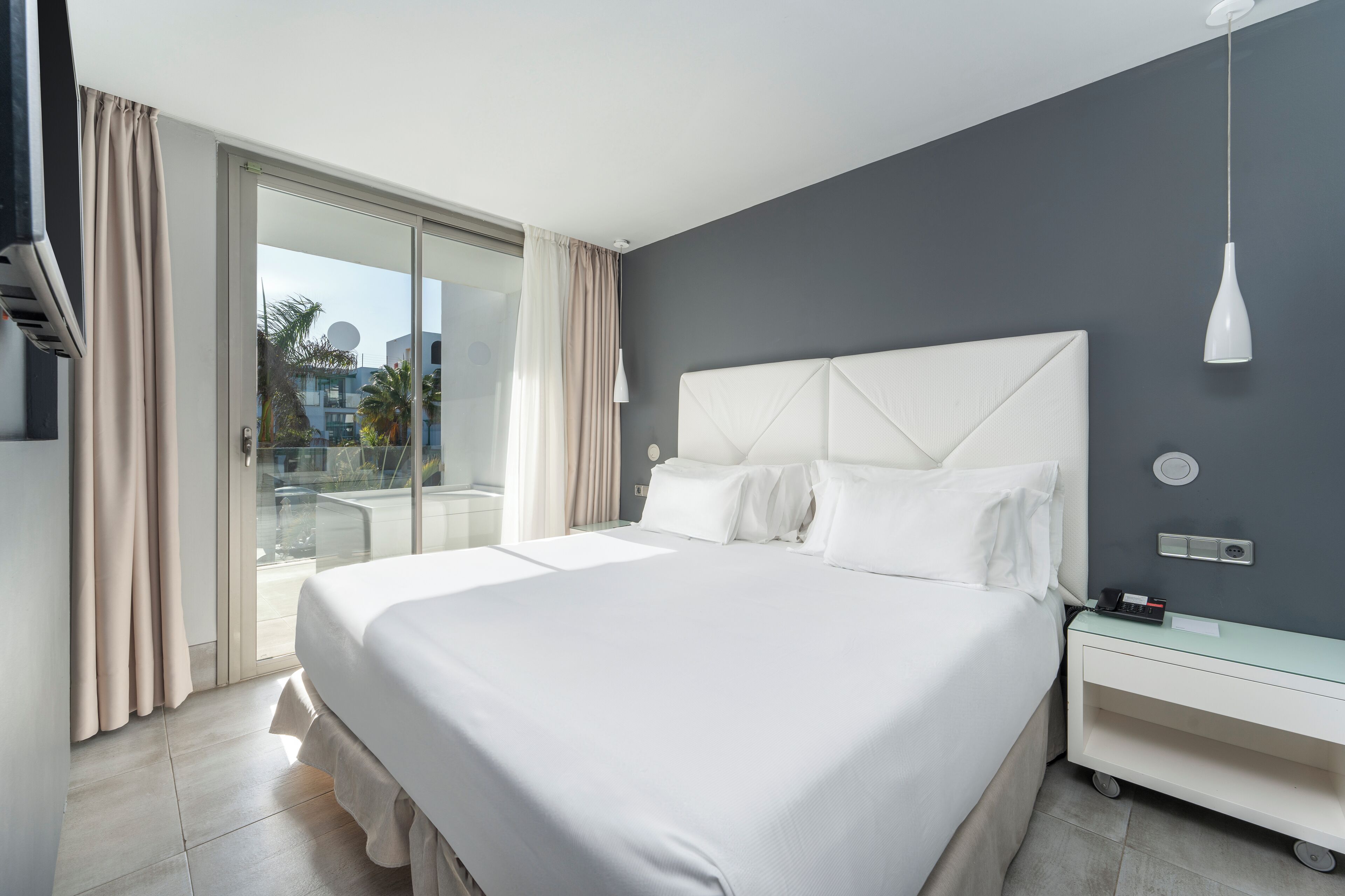 A modern hotel room with a large bed, a glass door leading to a balcony, and a white nightstand with a lamp.