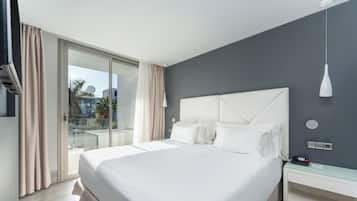 A modern hotel room with a large bed, a glass door leading to a balcony, and a white nightstand with a lamp.