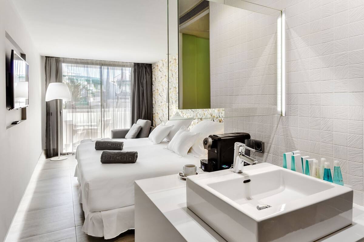A hotel room with a bed, a large mirror, a coffee machine, a sink, and toiletries.