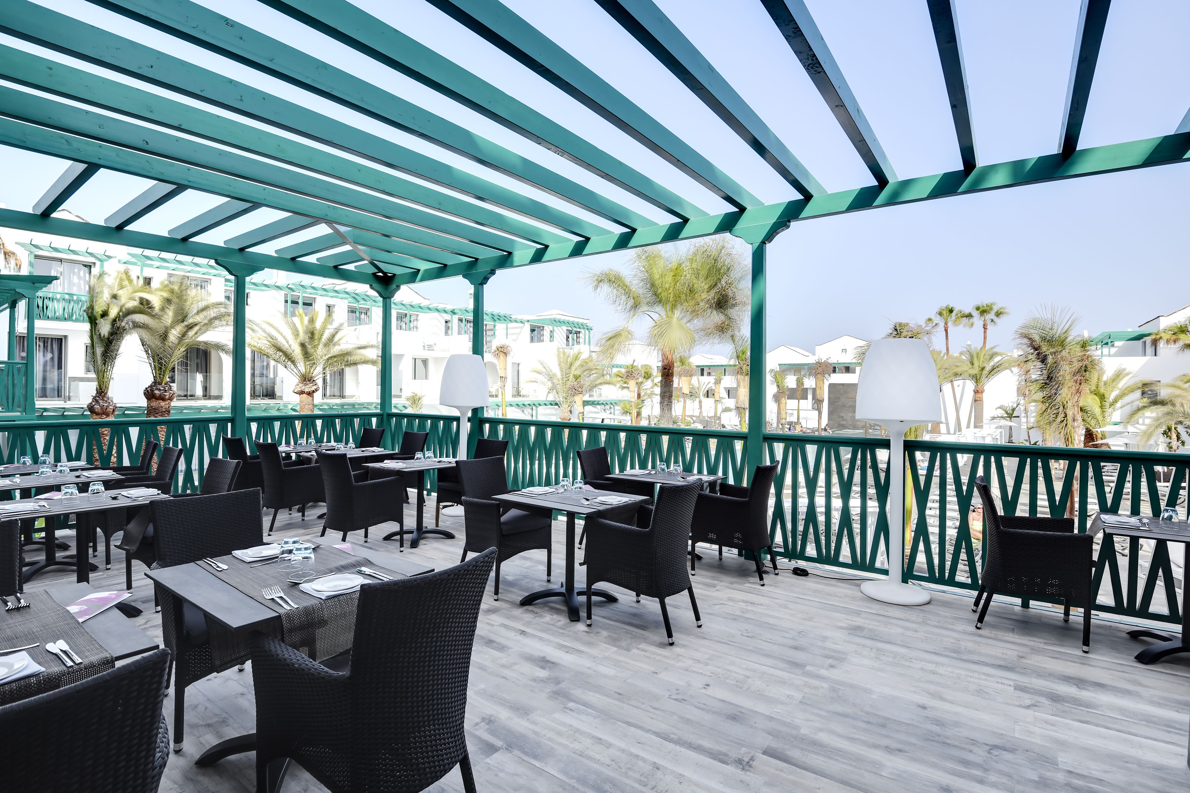An outdoor patio with tables and chairs, a green railing, and palm trees in the background.