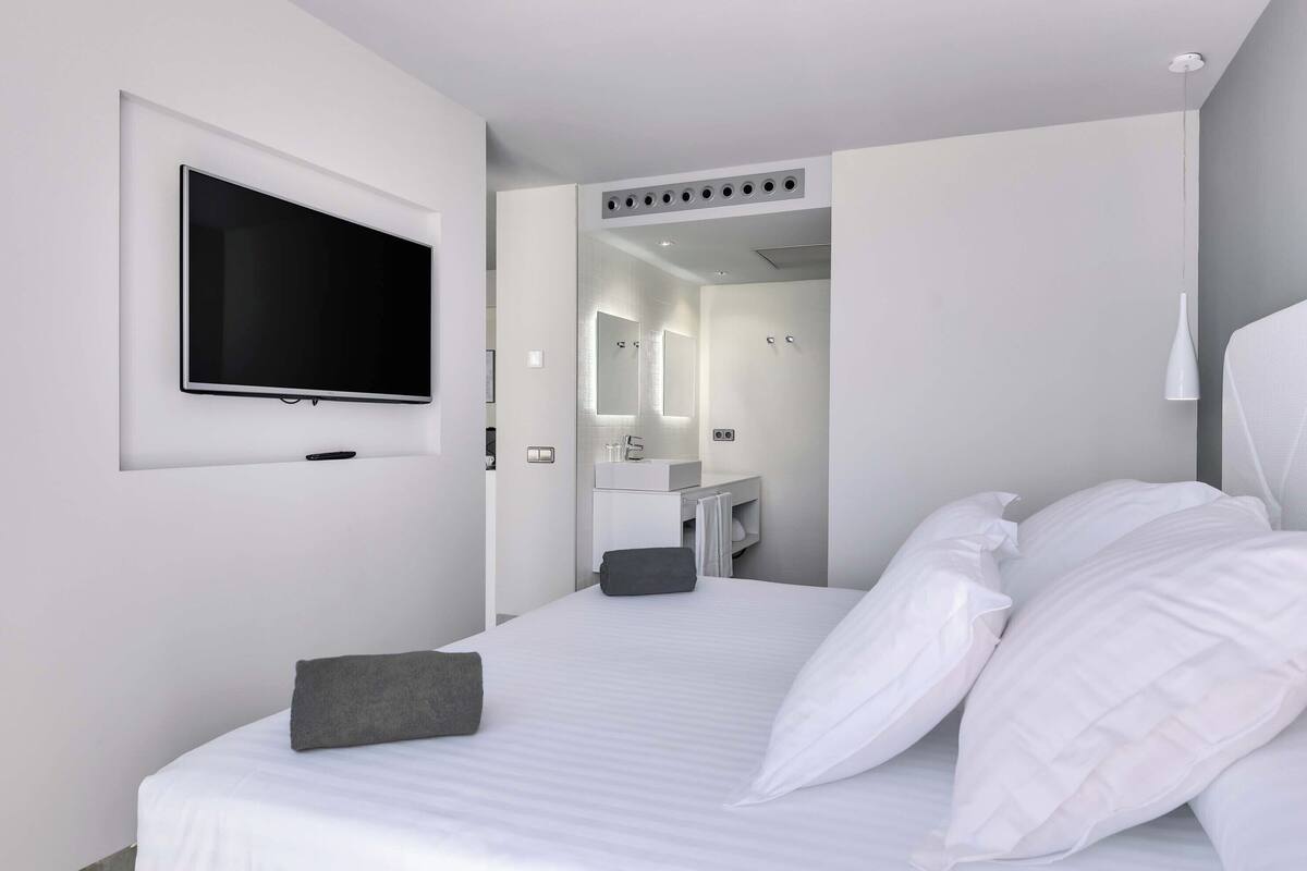 A modern hotel room with a large bed, a flat-screen TV mounted on the wall, a bathroom with a mirror and sink, and a walk-in closet.
