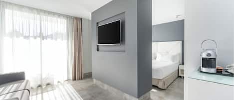 Une chambre d’hôtel moderne avec un téléviseur à écran plat fixé au mur, un lit avec une tête de lit blanche, une machine à café posée sur une table en verre et un canapé dans un coin.