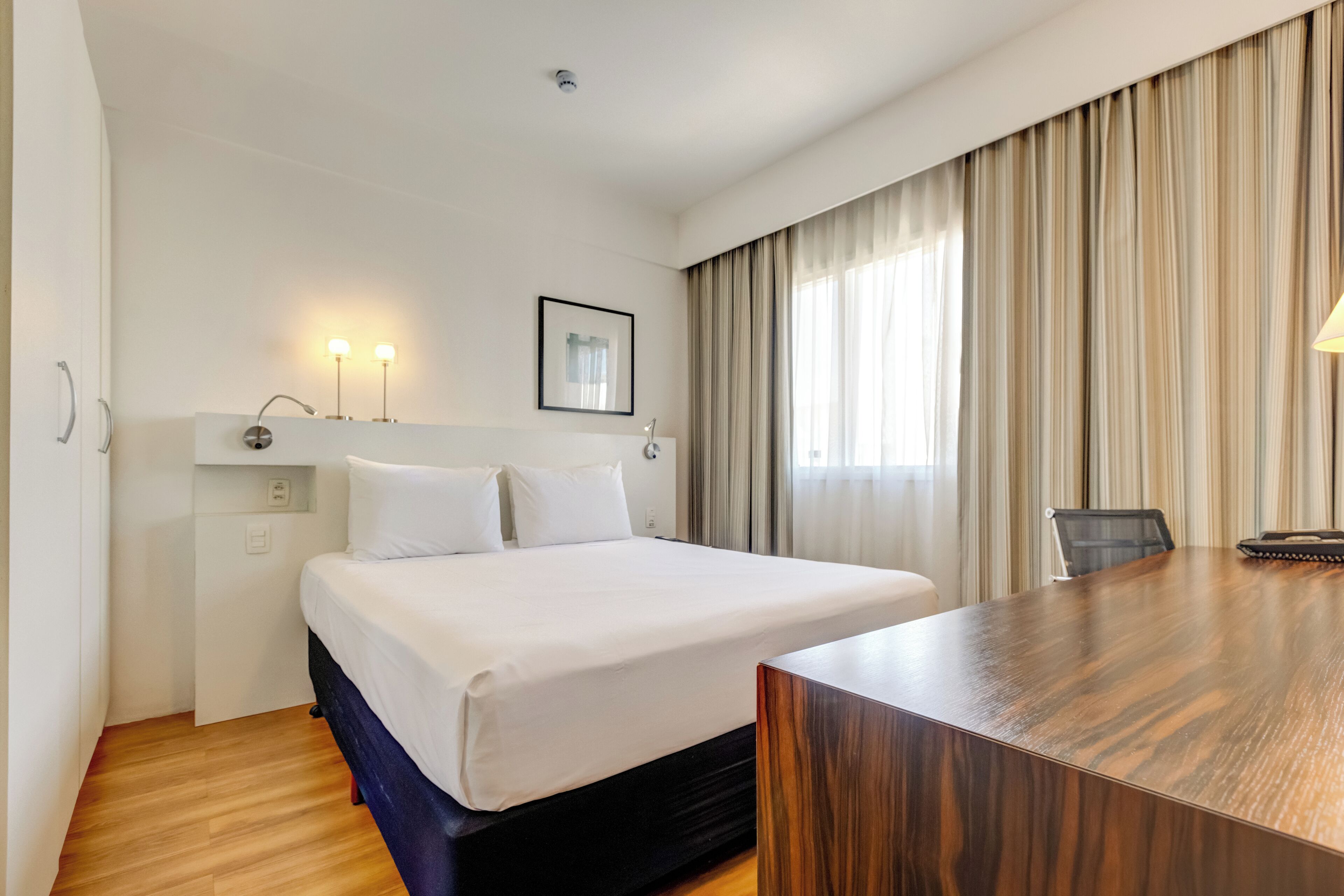 Habitación de hotel con una cama grande, un escritorio, una silla, un cuadro en la pared y una ventana con cortinas.