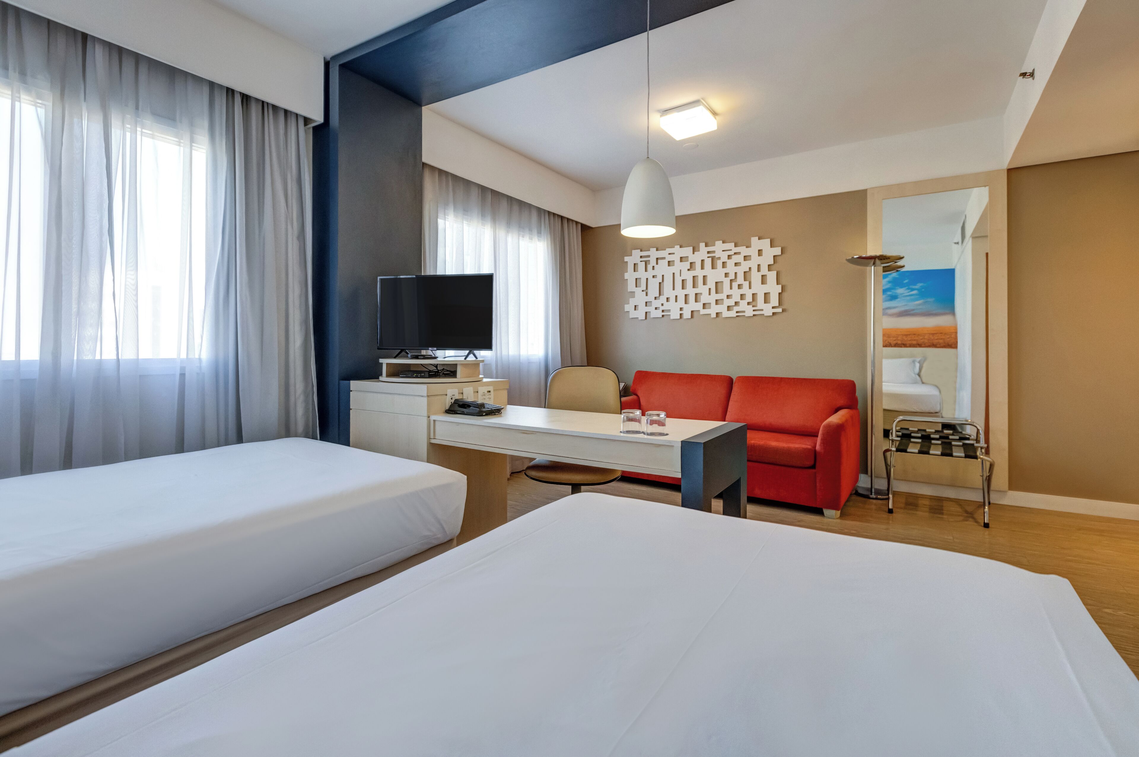 A hotel room with two beds, a desk with a TV, a red sofa, and a small table with glasses.