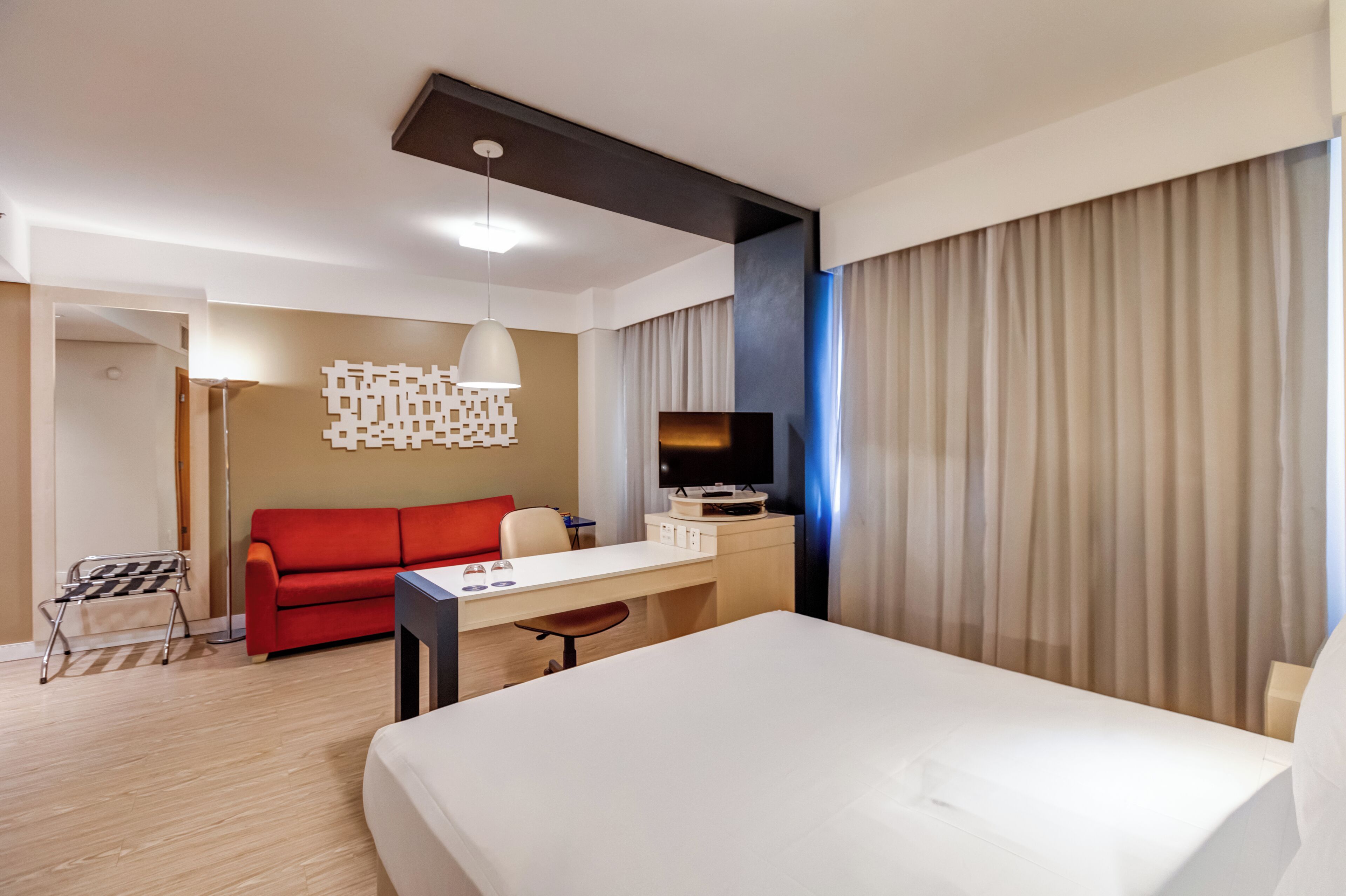 A hotel room with a bed, a desk with a TV, a red sofa, and a modern light fixture.